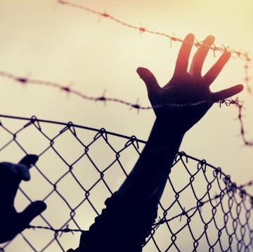 Silhouette of a hand reaching through a chain-link fence.