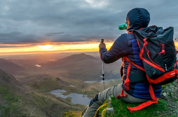 A hiker sits atop a mountain looking as the sun sets across a vast valley below.