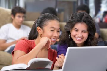 Two students smiling and using a laptop in a classroom.
