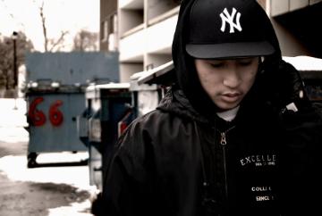A person in a dark jacket and cap looks down while walking outdoors.
