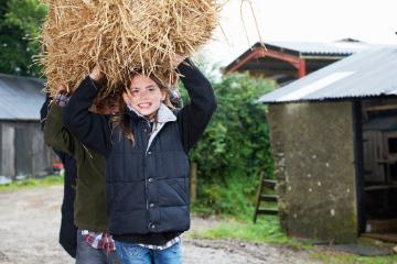 Child carrying a large bundle of straw outdoors on a farm. 