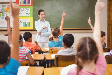 A classroom with students raising hands and a teacher at the chalkboard.