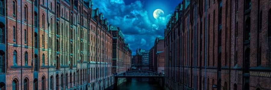 A full moon peaking out of clouds over city buildings with a river running through them and a bridge in the distance