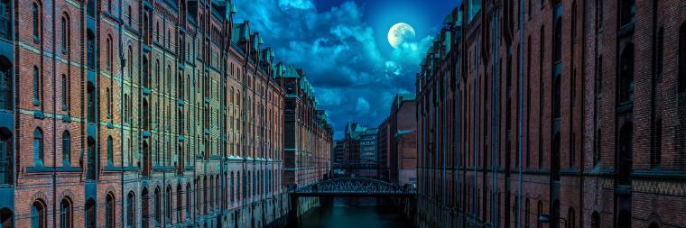 A full moon peaking out of clouds over city buildings with a river running through them and a bridge in the distance