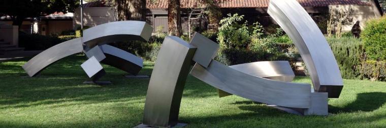 Curved metal sculpture in park with trees and benches.